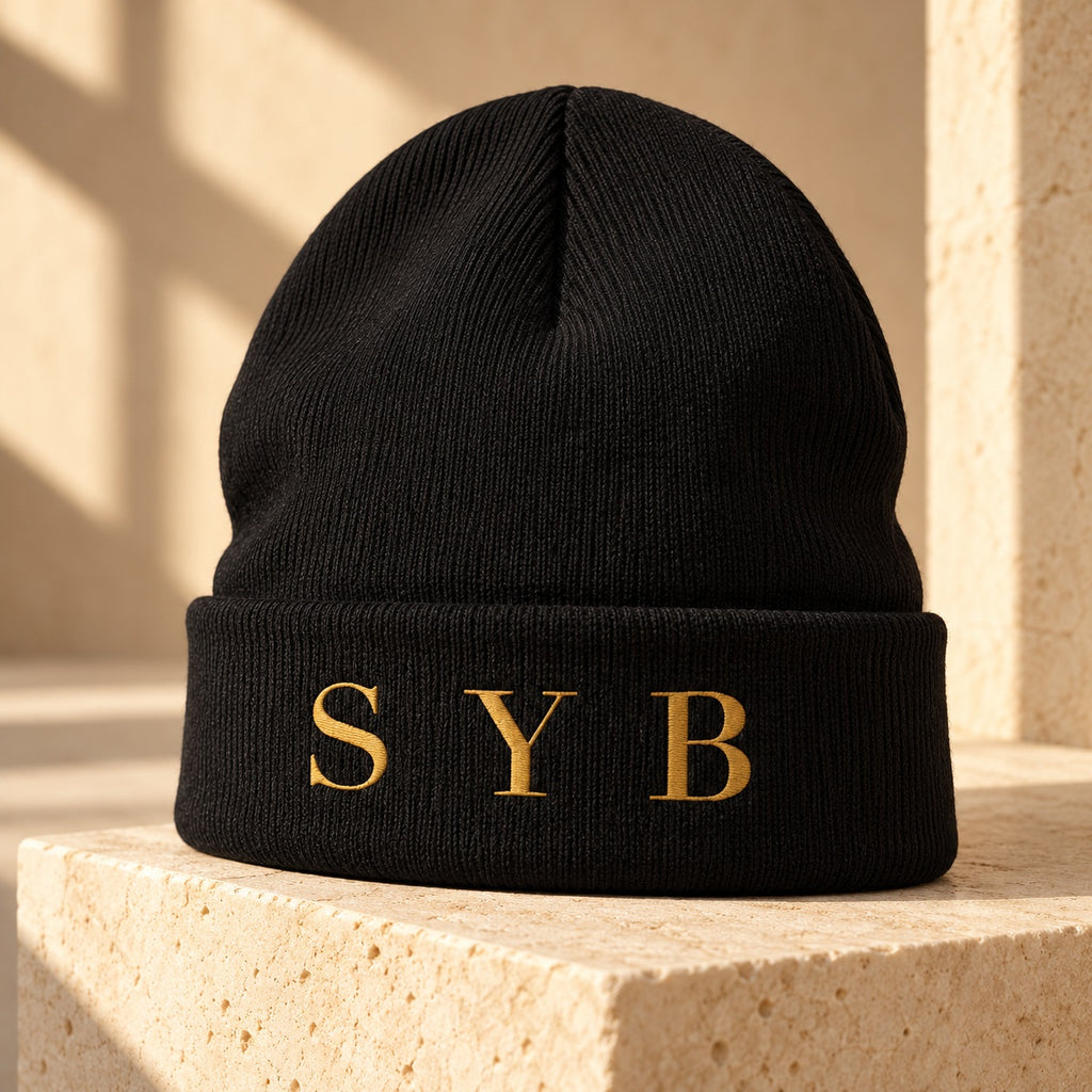 Black SYB Wordmark Beanie with Old Gold embroidery on limestone