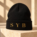 Black SYB Wordmark Beanie with Old Gold embroidery on limestone