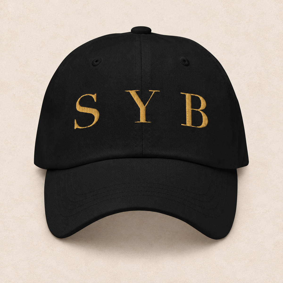 wordmark cap in black with original SYB gold artwork on a pale limestone studio background