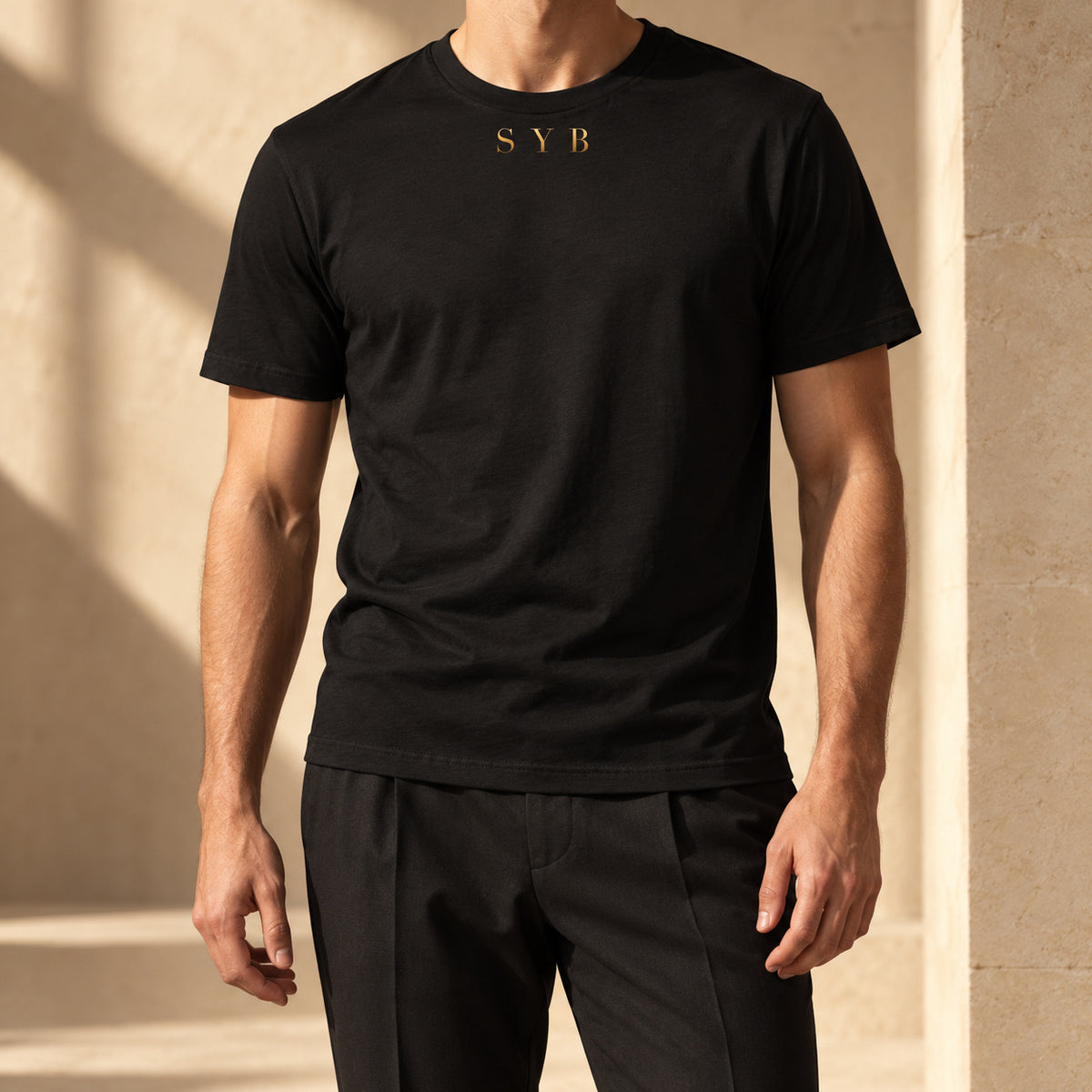 Wordmark Tee in black, styled with tailored trousers; gold-tone SYB front print in a warm limestone setting.